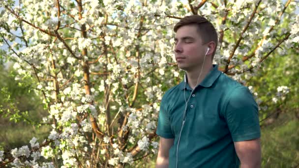 Jeune homme dans la nature avec des écouteurs dans les oreilles. Un homme écoute de la musique tout en se tenant contre un arbre à fleurs. — Video
