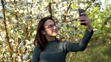 Genç bir kadın kendini bir elma ağacının arka planında fotoğraflıyor. Çiçek açan bahçede bir kız.