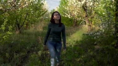 Genç bir kadın elma ağaçları arasında yürüyor. Bir kız çiçek açan bir bahçede yürüyor. Ön görünüm.