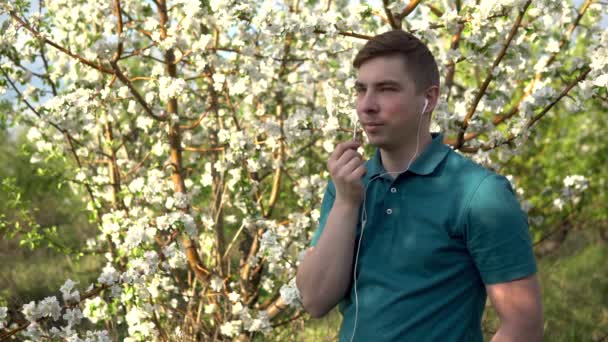 Joven en la naturaleza con auriculares. Un hombre está hablando por teléfono a través de auriculares mientras está de pie contra un árbol floreciente . — Vídeo de stock