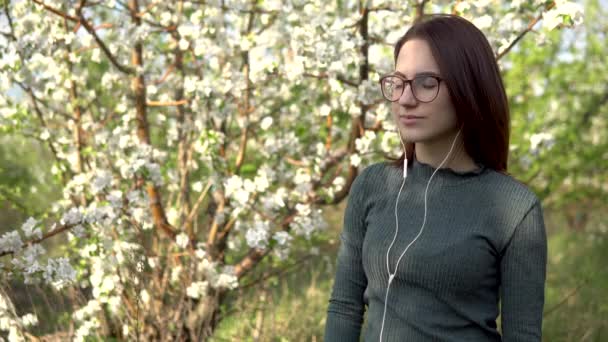 Mujer joven en la naturaleza con auriculares en los oídos. Una chica escucha música mientras está de pie contra un árbol floreciente . — Vídeo de stock