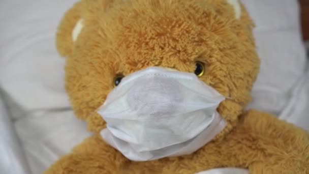 एक टेडी भालू एक चिकित्सा मुखौटा में बिस्तर पर पड़ा है। भालू बीमार हो गया . — स्टॉक वीडियो