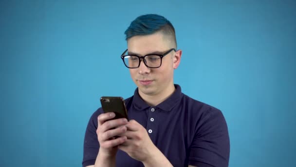Mavi saçlı, elinde telefon olan gözlüklü genç bir adam. Alternatif bir kişi mavi arka planda akıllı bir telefonla yazıyor.. — Stok video