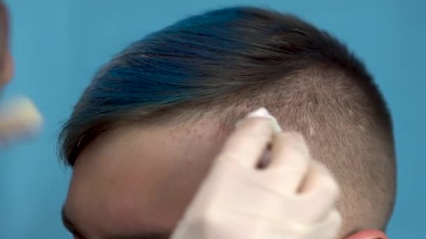 Egy kék hajú fiatalembert lemosnak az arcáról a festék miatt. Alternatív férfi egy szépségszalonban, kék alapon. — Stock videók