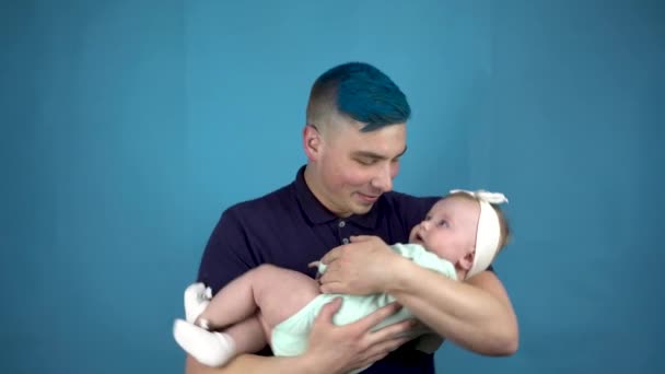 Um jovem pai com cabelo azul segura um bebê em seus braços. Pessoa alternativa embala uma criança em um fundo azul. — Vídeo de Stock
