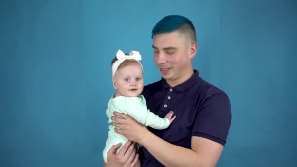 Młody ojciec z niebieskimi włosami trzyma dziecko w ramionach. Alternatywny mężczyzna z dzieckiem patrzy w kamerę na niebieskim tle. — Wideo stockowe