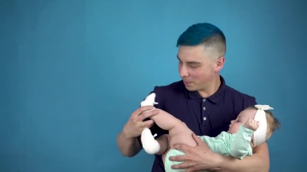 Ένας νεαρός πατέρας με μπλε μαλλιά κρατά ένα μωρό στην αγκαλιά του. Εναλλακτικός άνθρωπος βάζει μια κάλτσα σε ένα παιδί σε μπλε φόντο. — Αρχείο Βίντεο