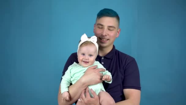 Mladý otec s modrými vlasy drží dítě v náručí. Alternativní muž s dítětem pohled na kameru na modrém pozadí. — Stock video