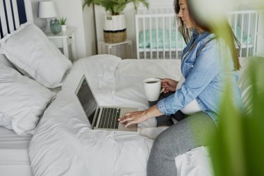Hamile kadın yatakta oturuyor ve bilgisayar kullanıyor.