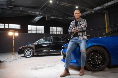 Kameraya gururla gülümseyen yakışıklı Afrikalı adam garajda spor arabasına yaslanıyor.