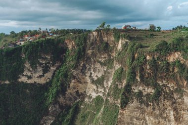 Üzerinde yeşil çimlerin ortasında evler ve arka planda bulutlar olan yüksek bir dağın fotoğrafı.