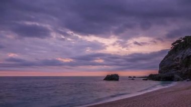 Uçurumlar ve sakin deniz arasında küçük bir yaz plajı. Bulutlu bir gökyüzünde şafak birçok renkte. Zaman aşımı
