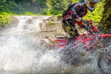 Ukraine, Vizhnitsa - August 25,2018: Summer day. A lot of splash of water hides an enduro motorcycle when an athlete overcomes a forest stream. Close-up clipart