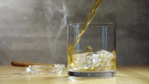 木製のテーブルの灰皿の中のハイボールガラスと葉巻の煙 ウイスキーの流れが氷の上に注ぐ スローモーション — ストック動画