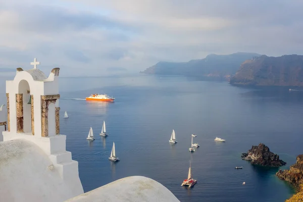 Yunanistan. Santorini 'de bulutlu bir gün. Oia 'da haçı olan bir Yunan kilisesinden yelkenli yatları ve yolcu gemisi olan bir körfeze.