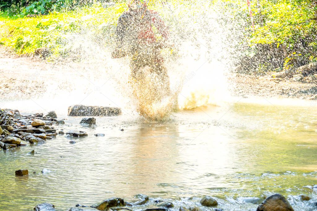 Sunny summer day. A lot of water spray hides athlete enduro when he overcomes a forest stream