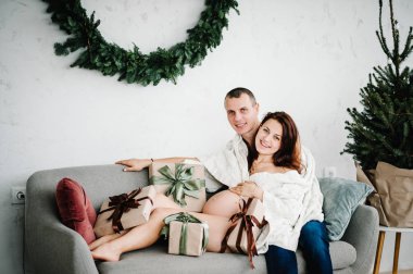 Hamile kadın ve erkek evde Noel ağacının yanında ekoseyle kaplanmış. Bir sürü hediye, aile. Mutlu yıllar ve mutlu Noeller. İç dekore edilmiş. Hamilelik, tatiller, insanlar ve beklenti konsepti.