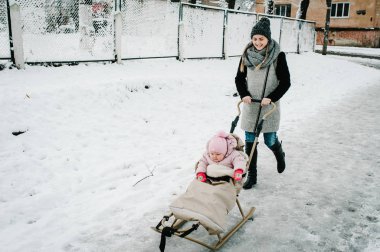 Genç ve mutlu bir anne, kışın arka planda bebek ve bir çocukla birlikte dışarı çıkar. Kapatın. Yolun yakınındaki bahçede mutlu bir aile ve şehrin sokaklarında binalar. Bebek..