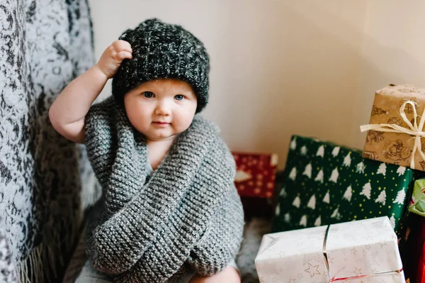 Portrait of little girl in warm knitted scarf sitting in room on gifts background, festive birthday concept