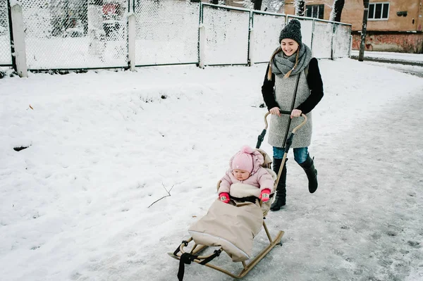A happy young mother walks with baby and a children sled outdoors on the background  winter. Close up. Happy family in the yard near the road and buildings on the streets of the city. Infant.