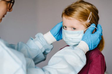 Hemşire, koruyucu giysili doktor, çocuk için yüzünde tıbbi maske var. Covid-19 enfeksiyonuna karşı önleyici önlemler. Gribe karşı korunma kavramı, korona virüsü salgını.
