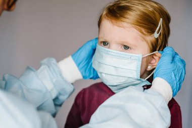 Coronavirus. Hemşire, koruyucu kıyafetli doktor çocuk için tıbbi maske takıyor. Covid-19 enfeksiyonuna karşı önleyici önlemler. Gribe karşı korunma kavramı, korona virüsü salgını.