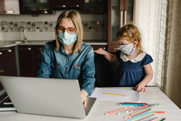 Coronavirus. Karantinada anne ve kızı koruyucu maske takıyor. Evde kalıp bebeğe bakarken dizüstü bilgisayarda çalışan bir kadın. Serbest bir iş. Çocuk gürültü yapıyor, oynuyor ve anneyi rahatsız ediyor..