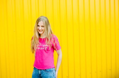 Pembe gömlekli ve kot pantolonlu uzun sarı saçlı güzel atletik genç bayan. Sarı arka plandaki garaj duvarında poz vermek ve gülümsemek. Eller kemerde. Mesaj için yer. Yakın plan. Gülümsüyor, poz veriyor