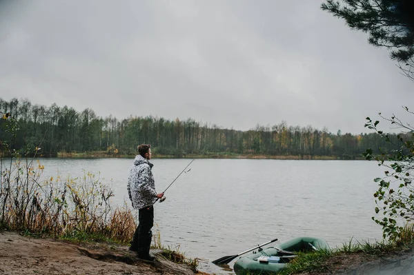 Fisherman with a fishing rod near an inflatable boat, with boxes with fishing tackles, is standing on the water, pond, lake, near the shore on the background of tree, nature. Fishing background. boat with oars.