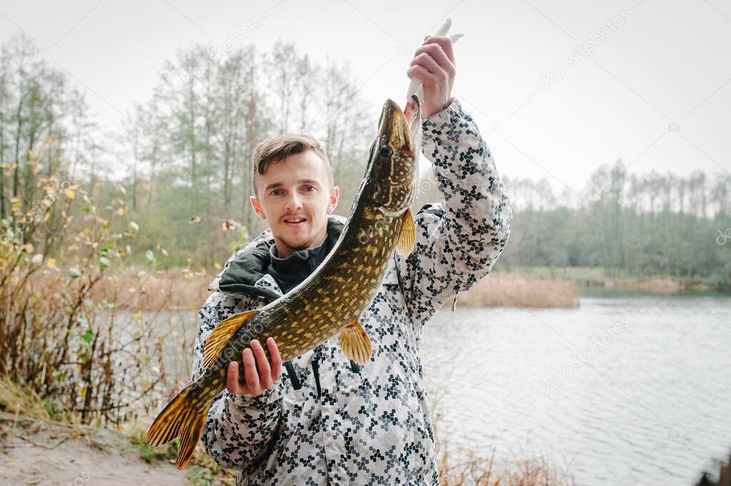 Happy amazed cheerful young fisherman hold a big fish pike near the boat on a background of lake and nature. Fishing background. Good catch. Trophy fish. angler. close up. headshot.