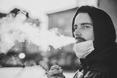 Sigara içiyorum. COVID-19 salgını sırasında maskeli bir adam sokakta sigara içiyor. Sigara içmek akciğer kanseri ve diğer hastalıklara neden olur. Sigara içmenin tehlikeleri. Siyah beyaz fotoğraf. Coronavirüs.