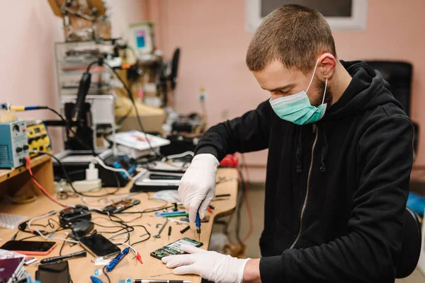 The technician repairing the smartphone motherboard in the lab. Concept of mobile phone, electronic, repairing, upgrade, technology.  Coronavirus. Man working, wearing protective mask in workshop.