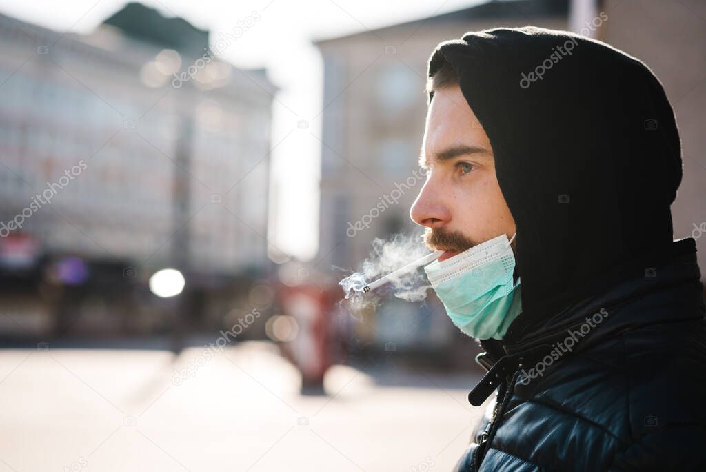 Smoking. Closeup man with mask during COVID-19 pandemic smoking a cigarette at the street. Smoking causes lung cancer and other diseases. The dangers and harm of smoking. Coronavirus.