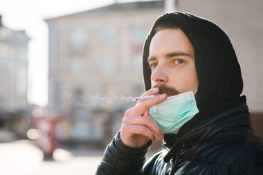 Sigara içiyorum. COVID-19 salgını sırasında maskeli bir adam sokakta sigara içiyor. Sigara içmek akciğer kanseri ve diğer hastalıklara neden olur. Sigara içmenin tehlikeleri ve zararları. Coronavirüs.