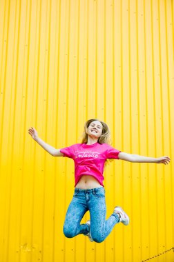 Pembe gömlekli ve kot pantolonlu uzun sarı saçlı güzel atletik genç bayan. Sarı arka plandaki garajın duvarında poz verip gülümsüyor. Mesaj için yer. Sıçrıyor, uçuyor. Rüzgâr saçı sürükler