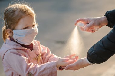 Coronavirus. Kadın, sokakta koruyucu maske takmış bir çocuğa sprey dezenfektan sıkar. Covid-19 enfeksiyonuna karşı önleyici önlemler. Anti bakteriyel yıkama spreyi. Hastalık koruması.