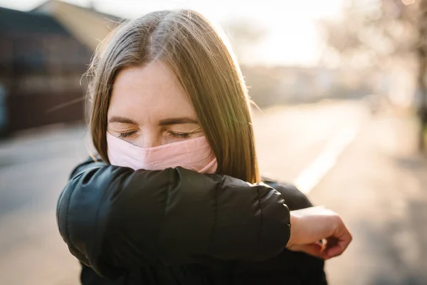 Girl with mask during COVID-19 pandemic coughing at the street. The dangers of Coronavirus. Risk of spreading infection. Covering nose and mouth, sneezing flexed elbow. Woman cough in arm prevention.
