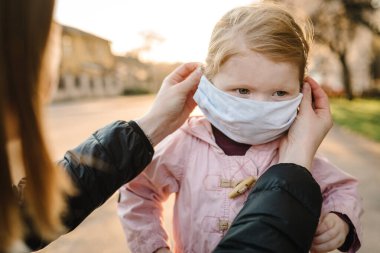 Coronavirus ve Hava kirliliği konsepti. Maske takan küçük bir kız ve anne sokakta yürüyor. Annem maskeli çocuğu düzeltiyor. Salgın virüs belirtileri. Dışarıda çocuklu bir aile. Hastalık koruması.