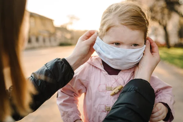 Coronavirus ve Hava kirliliği konsepti. Maske takan küçük bir kız ve anne sokakta yürüyor. Annem maskeli çocuğu düzeltiyor. Salgın virüs belirtileri. Dışarıda çocuklu bir aile. Hastalık koruması.