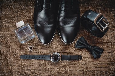 Siyah ayakkabılar, siyah kuşak, siyah saat, siyah kelebek, kol düğmeleri ve parfüm kahverengi bir arka planda.