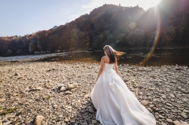 Beyaz elbiseli gelin nehir taşının üzerinde rüzgar saçlarını savuruyor ve güneş parlıyor.