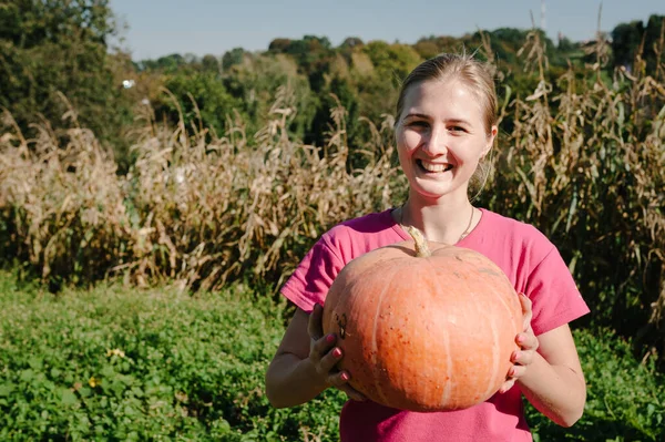 Portrait of cheerful woman with pumpkin in hands over background field, picking out on a farm at sunset. Autumn harvest. Vegetable garden. Growing pumpkins. Celebrating happy Thanksgiving day.