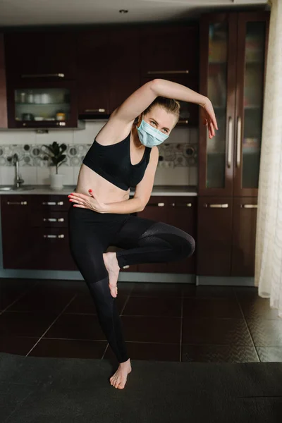 Girl in protective mask, sportswear on floor on mat at home. Woman are practicing yoga at home. Female doing morning work-out. Coronavirus. Stay Home. Sport at home. Calmness concept. Fitness workout.