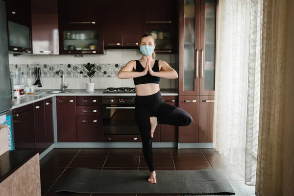 Girl in protective mask, sportswear on floor on mat at home. Woman are practicing yoga at home. Female doing morning work-out. Coronavirus. Stay Home. Sport at home. Calmness concept. Fitness workout.