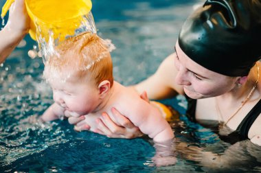 Genç anne, havuzda mutlu küçük kız. Çocuğa yüzmeyi öğretiyor. Suda yüzmenin ilk gününün tadını çıkar. Anne çocuğu tutuyor. Egzersiz yapıyorum. Elleri bebeğin kafasına bir bardak su döker.