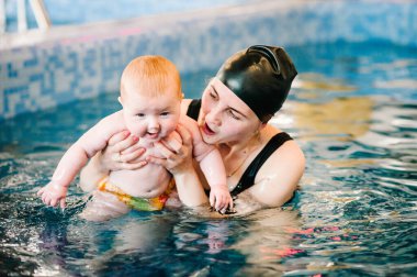 Kürek havuzuna bebek dalışı. Genç anne, yüzme öğretmeni ve bilardodaki mutlu küçük kız. Küçük çocuğa yüzmeyi öğret. Suda yüzmenin ilk gününün tadını çıkar. Anne bebeği tutuyor ve dalıyor.