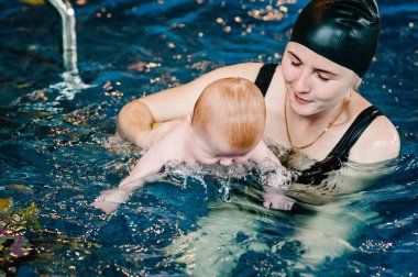Kürek havuzuna bebek dalışı. Genç anne, yüzme öğretmeni ve bilardodaki mutlu küçük kız. Küçük çocuğa yüzmeyi öğret. Suda yüzmenin ilk gününün tadını çıkar. Anne bebeği tutuyor ve dalıyor.