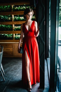 Mezuniyet için kırmızı elbiseli, çantalı esmer kız. Panoramik pencerenin yanında. Rafların arka planında yeşil yapraklar ve esrar var. Parti zamanı. Uzun elbiseli kadın restoranda bekliyor..