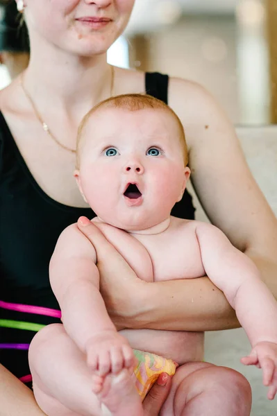 mom holding baby girl 6 months, a baby in her arms at the pool. The first swimming lesson. Prepare to dive. Surprised. with an open mouth. red-head hair. close-up. infant. Keep your feet in hands