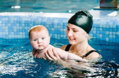 Genç anne, yüzme öğretmeni ve havuzdaki mutlu küçük kız. Çocuğa yüzmeyi öğretiyor. Suda yüzmenin ilk gününün tadını çıkar. Anne dalışa hazırlanan çocuğun elini tutuyor. Egzersiz yapmak